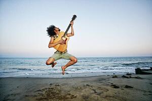 Fotografija Mixed Race man playing guitar and jumping at beach, Peathegee Inc, (40 x 26.7 cm)